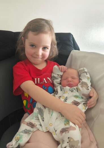 A young girl with light brown hair and a red shirt smiles while lovingly holding a newborn baby dressed in an animal-patterned onesie, sitting together indoors.