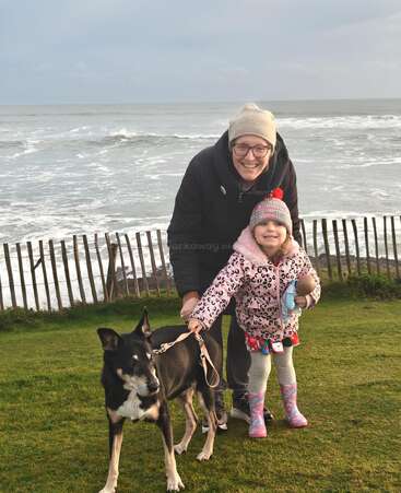 A smiling adult and child stand together on green grass by the sea, holding a dog on a leash. The child holds a doll, both warmly dressed.