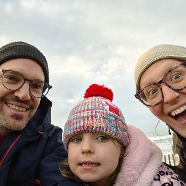 A cheerful family selfie outdoors features two smiling adults wearing glasses and hats, and a bundled-up child in a colorful knitted hat and pink coat. Cloudy sky.