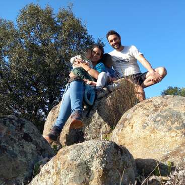 A happy family of three sits atop large rocks outdoors, surrounded by greenery and trees. The sky is clear and blue, and everyone is smiling joyfully.