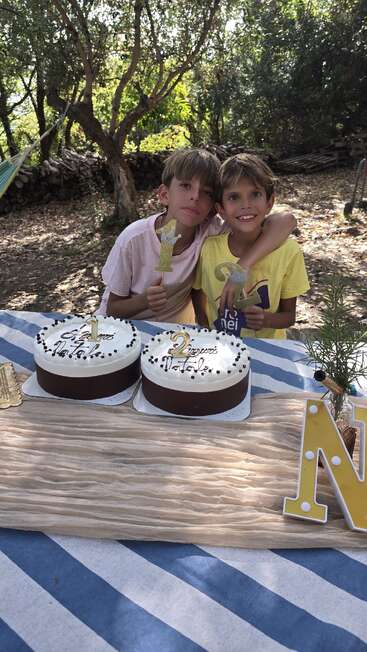Dois meninos estão sentados ao ar livre em uma mesa, sorrindo e abraçados. Dois bolos de aniversário com velas e decorações estão na frente deles, comemorando juntos.