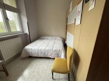 Ein kleines Schlafzimmer mit einem Doppelbett, einem gelben Stuhl, Fliesenboden, einem großen Fenster, einem Heizkörper und einem Wandregal, das mit verschiedenen Bildern oder Postern über dem Bett dekoriert ist.