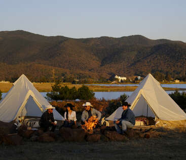 Four people wearing hats sit around a campfire between two tents by a lake, surrounded by mountains and trees, enjoying an outdoor camping experience at sunset.
