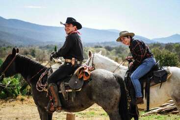 Two people dressed as cowboys sit smiling on horseback in a scenic outdoor setting, with mountains and trees in the background, enjoying a sunny day.