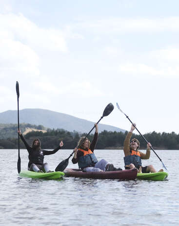 Three people in life jackets sit in kayaks on a calm lake, smiling, raising their paddles triumphantly, with mountains and trees in the background. Outdoor adventure fun.