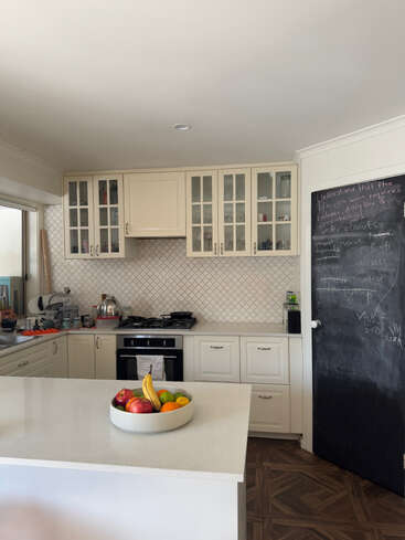 Das Bild zeigt eine moderne Küche mit weißen Schränken, einer weißen Arbeitsplatte und einer Wandtafel, auf der eine Schale mit Obst steht und im Hintergrund ein Herd.