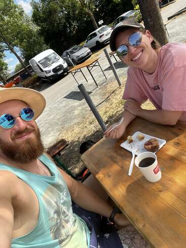 Two people are sitting at a wooden outdoor table, enjoying coffee and cake. Both are smiling, wearing sunglasses, casual summer clothes, surrounded by parked cars and trees.