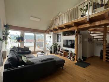 This cozy living room features a sectional sofa, large windows, indoor plants, wooden floors, a mezzanine, TV, open staircase, and modern, inviting decor with natural light.