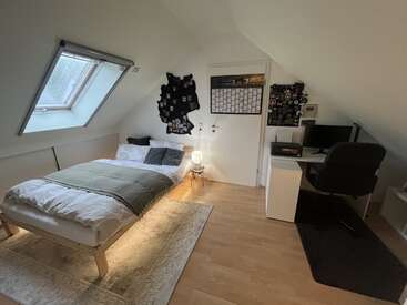 A cozy attic bedroom with a slanted ceiling features a bed, desk, computer, patches on the wall, a window, soft lighting, and minimalist decor.
