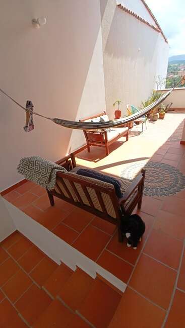 Sunny terrace with terracotta tiles, wooden chairs, hammock, potted plants, and a mosaic design. A black and white cat sits in the shade near the stairs.