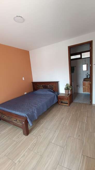 A simple room with a single bed, wooden headboard, blue quilt, nightstand with a plant, orange accent wall, tiled floor, and an attached bathroom.