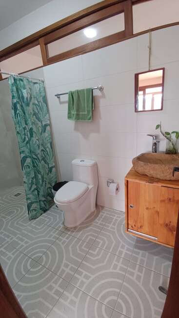 This bathroom features a leafy shower curtain, green towel, modern toilet, wooden vanity with a stone sink, geometric patterned tiles, and a hanging mirror with plants.