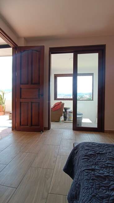 This image shows a cozy room with wooden doors, tiled floors, and a bed with a dark quilt. A sunlit sitting area and balcony are visible.