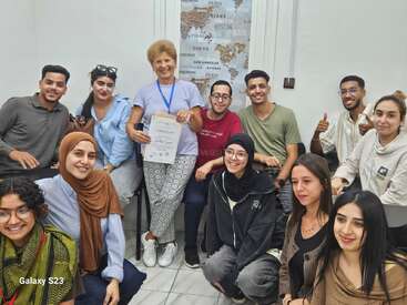 A group of smiling students surrounds a woman holding a certificate. They are in a classroom with a world map, looking cheerful and proud, celebrating achievement.