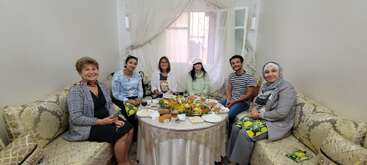 Seven people are sitting around a round table, sharing a meal in a cozy, well-lit living room. They are smiling, enjoying food, and spending time together.