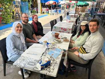 Five people are sitting together at an outdoor restaurant table, smiling. There’s food and drinks on the table, menus visible, and colorful umbrellas in the background.