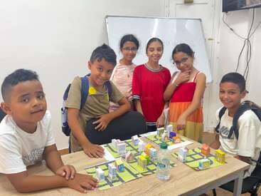 Six smiling children gather around a table with a miniature city model. Behind them is a whiteboard, suggesting a classroom or educational environment. They look happy.