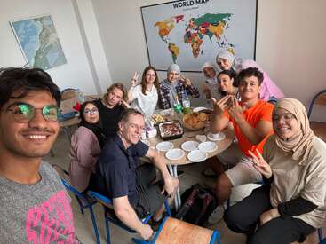 A group of diverse people happily sit around a table with food, posing for a selfie in a classroom. A world map hangs on the wall.