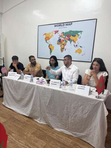 Five people sit at a table covered with a white cloth, discussing or presenting, with nameplates and flags. A colorful world map hangs behind them.