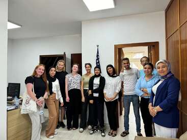 A group of eleven people, men and women, pose together in an office. An American flag stands in the background. They appear friendly and diverse.