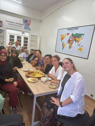 A diverse group of people enjoys a meal together in a classroom. There’s a world map on the wall and a “Learn Locally, Speak Globally” sign.
