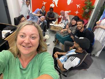 A group of people happily gather in a cozy room, smiling and posing for a selfie. Snacks and drinks are on the table. The atmosphere is friendly.