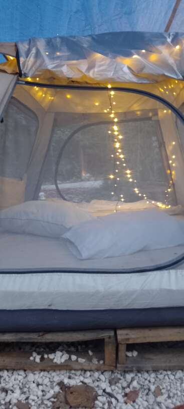 Una acogedora tienda montada sobre palés de madera, con una cama con almohadas y colchón blancos, adornada con luces de cuerda, crea un ambiente cálido y acogedor.