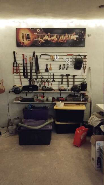 Una pared de taller bien organizada con diversas herramientas colgadas de forma ordenada, estantes con cascos y recipientes, contenedores de almacenamiento debajo, arte con guitarras y luces de cadena arriba para crear ambiente.
