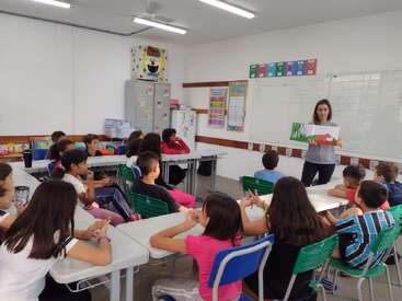 Un profesor está de pie al frente del aula, con un dibujo en la mano, dirigiéndose a los alumnos, que están sentados en sus pupitres y le escuchan con atención. El aula es luminosa, colorida y está bien ordenada.