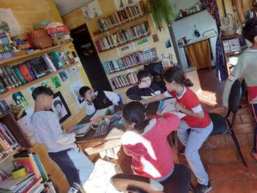Varios niños están sentados dibujando alrededor de una mesa de madera en una acogedora sala bañada por la luz del sol, llena de estanterías, material artístico y libros de colores, lo que crea un ambiente muy animado.