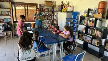 Una profesora está leyendo un libro ilustrado a un grupo de niños en un aula llena de estanterías, pupitres y material didáctico, lo que crea un ambiente animado.