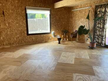 Una habitación espaciosa y minimalista con suelos y paredes de madera, una gran ventana, una planta en maceta, dos sillas, mesitas, luz natural y una cortina decorativa. Ambiente acogedor y tranquilo.