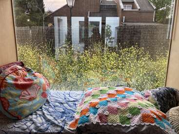 Un acogedor asiento junto a la ventana con coloridos cojines y un almohadón de ganchillo da a un jardín de flores amarillas, con casas y reflejos visibles a través del cristal.