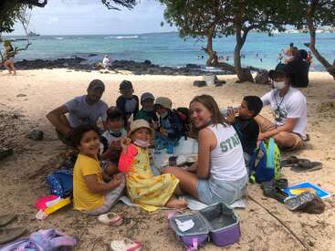 Um grupo de crianças e adultos está sentado em um piquenique na praia, sorrindo à sombra das árvores, com o mar e as pessoas nadando ao fundo.