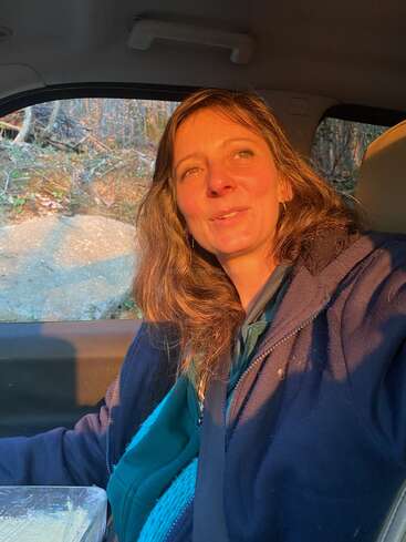 Una mujer está sentada en un coche, bañada por la cálida luz del sol, con una chaqueta azul y una suave sonrisa. A través de la ventanilla se ven árboles y la tierra.