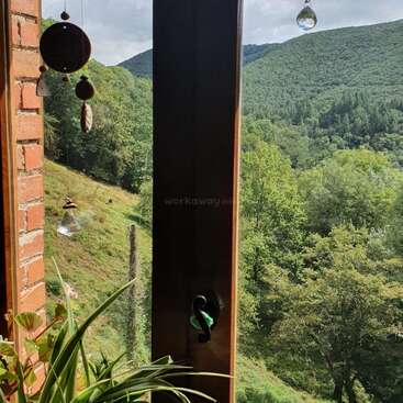 La imagen representa una serena ventana con vistas a una frondosa ladera boscosa, con una pared de ladrillo y campanillas de viento colgantes a la izquierda.