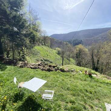 La imagen representa una serena ladera cubierta de hierba y árboles, con una mesa y sillas blancas, un cubo y un muro de piedra, sobre un pintoresco fondo montañoso.