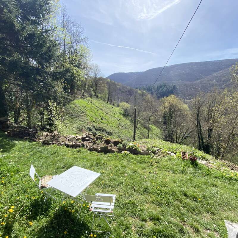 La imagen representa una serena ladera cubierta de hierba y árboles, con una mesa y sillas blancas, un cubo y un muro de piedra, sobre un pintoresco fondo montañoso.