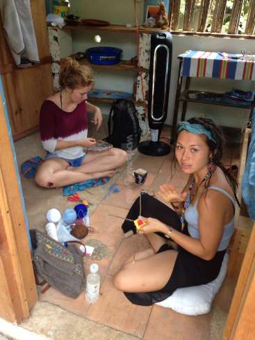 La imagen muestra a dos mujeres sentadas en el suelo de una pequeña habitación, rodeadas de material de manualidades, dedicadas a una actividad creativa.