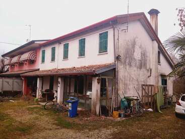 Esta imagen muestra una casa antigua y desgastada de dos pisos con techo rojo, ventanas con rejas, bicicletas dispersas, un contenedor de basura azul y varios objetos al aire libre alrededor.