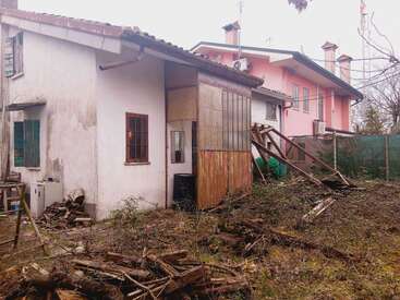 La imagen muestra una casa vieja y deteriorada con la pintura descascarillada junto a una casa más nueva y rosa. El jardín está cubierto de maleza y lleno de escombros y trozos de madera.