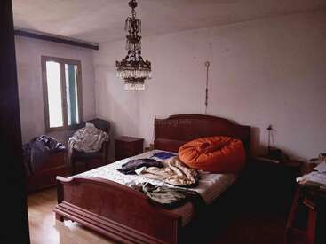 Una habitación con poca luz, con una cama de madera, ropa desordenada esparcida por todas partes, un puf naranja, muebles sencillos, una lámpara de araña y una ventana abierta.