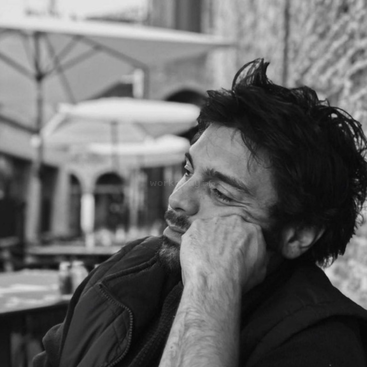 Un hombre está sentado en la terraza de un café, con la cabeza apoyada en la mano, mirando pensativo a lo lejos. La imagen es en blanco y negro, lo que evoca contemplación.