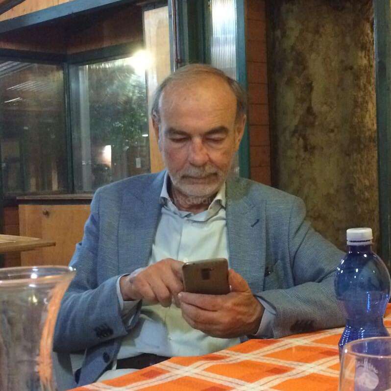 A imagem mostra um homem idoso sentado em uma mesa, olhando para o telefone, com uma garrafa de água e um copo por perto, em um ambiente rústico.