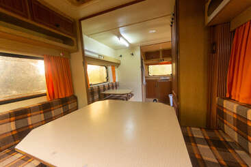 Esta imagen muestra el acogedor interior de una autocaravana clásica, con asientos a cuadros, cortinas naranjas, una mesa de comedor, armarios de madera, ventanas y una pequeña cocina americana.
