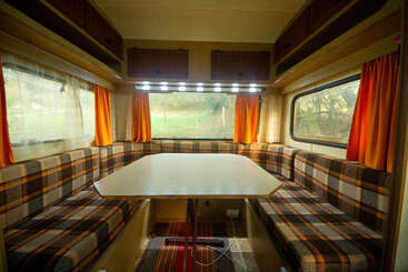 El acogedor interior de esta autocaravana cuenta con un banco a cuadros alrededor de una mesa rectangular, cortinas naranjas, armarios superiores y amplios ventanales que ofrecen vistas panorámicas y luz natural.
