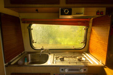 Esta imagen muestra la acogedora cocina de una autocaravana, con un fregadero, una cocina de dos fuegos y la luz del sol entrando por la ventana, lo que crea un ambiente cálido y acogedor.