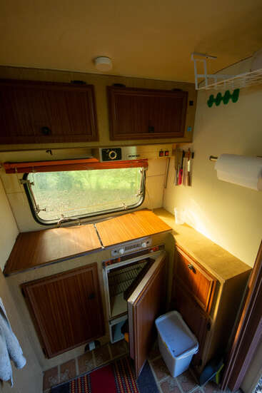 Esta imagen muestra una cocina compacta de autocaravana con armarios de madera, un horno pequeño, una nevera abierta, utensilios, un portarrollos, una ventana, una encimera y un cubo de basura.