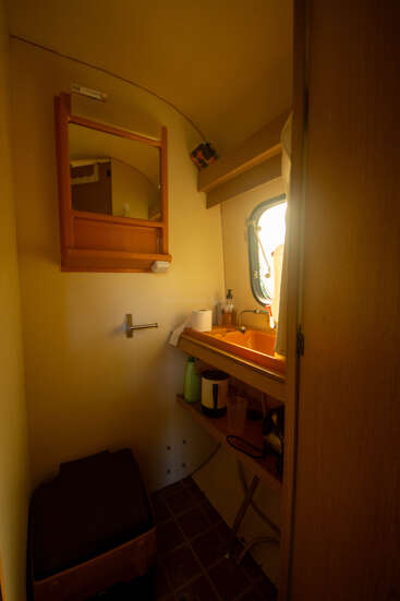 Esta imagen muestra un cuarto de baño compacto con un espejo, un lavabo pequeño debajo de una ventana, estantes con artículos de aseo, un inodoro y una toalla colgada cerca. Iluminación acogedora.