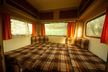 El acogedor interior de esta autocaravana cuenta con una gran cama con diseño a cuadros, cortinas naranjas y amplios ventanales que permiten que la cálida luz del sol ilumine este espacio acogedor de estilo retro.